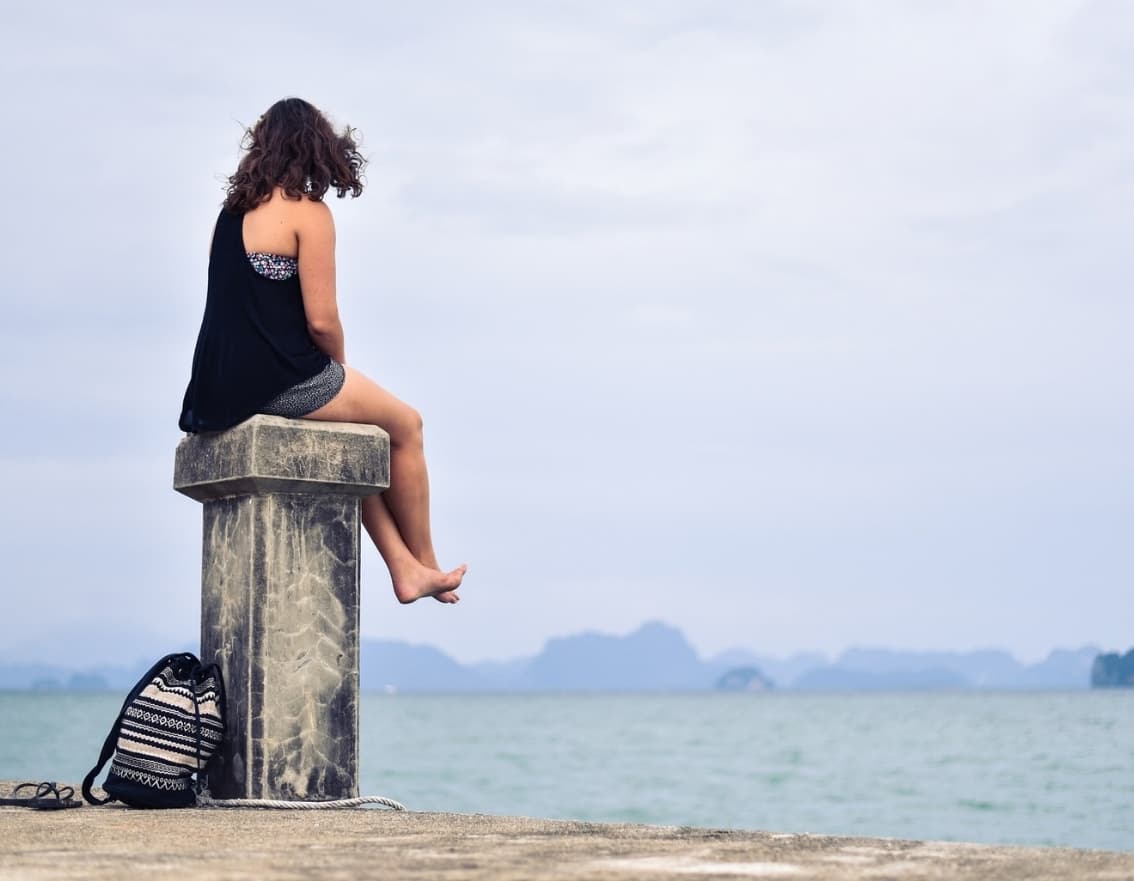 A woman sits alone on a harbour post, her back to the camera, looking out over calm water toward a distant horizon.