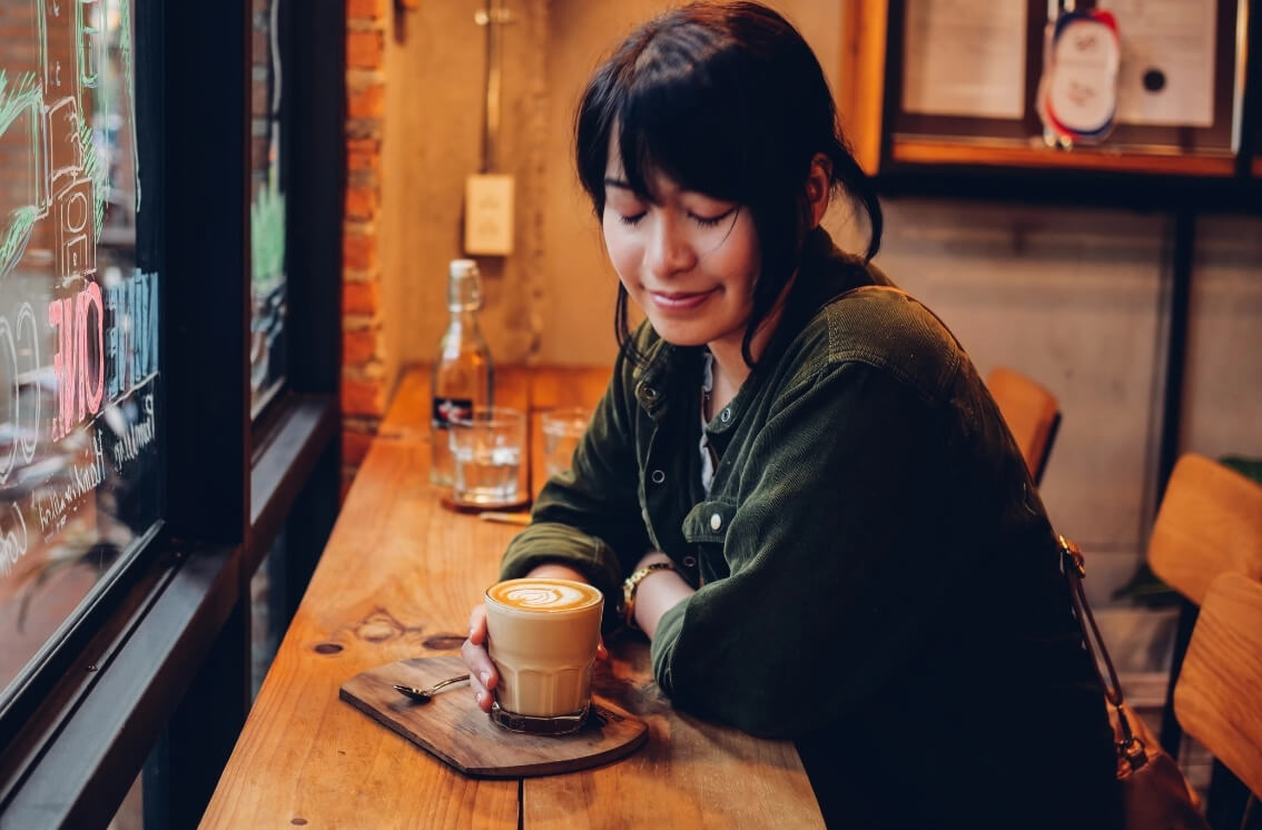 A woman sits alone in a warm cafe, hands around a coffee cup, lost in her own thoughts.