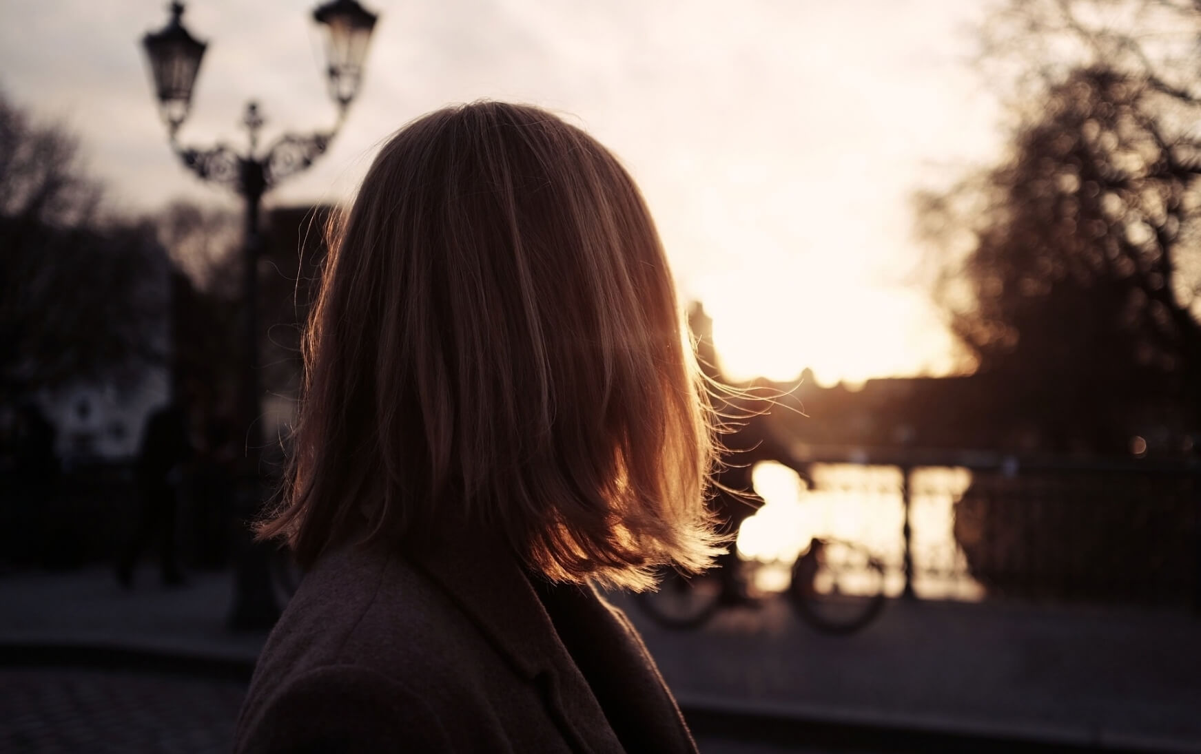 A woman stands alone on a city bridge at golden hour, her back to the camera, looking toward the light