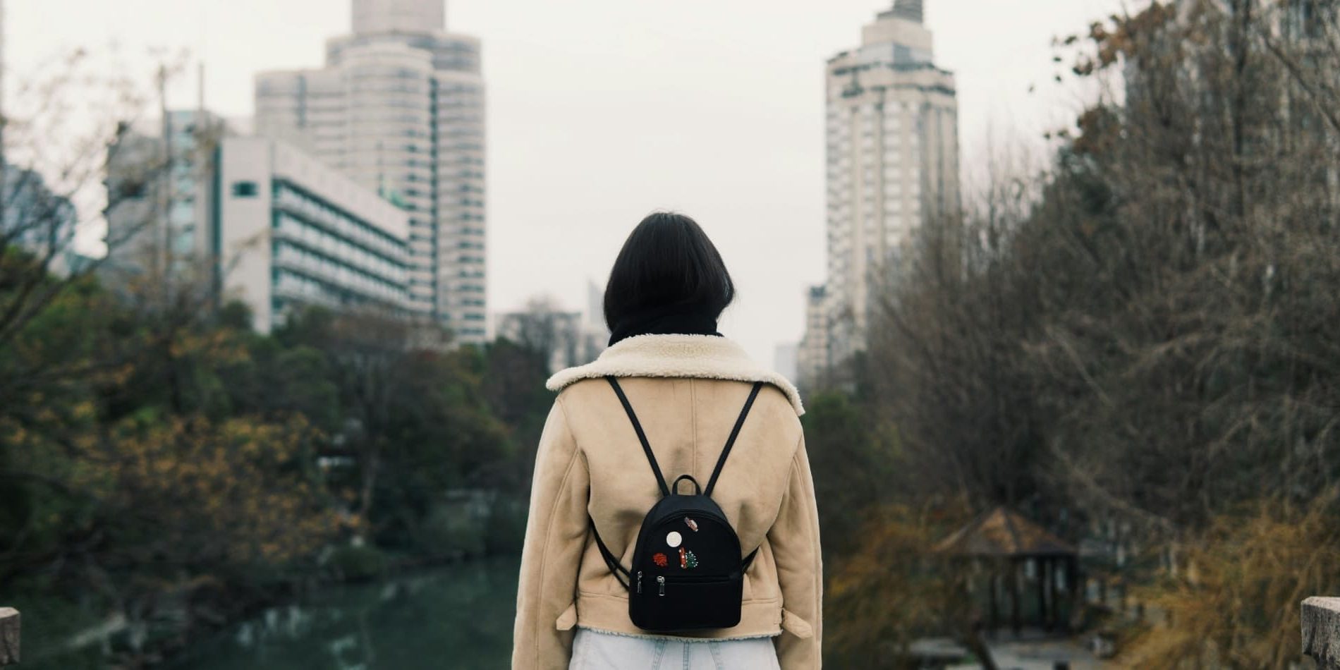 A woman stands alone on a bridge in an Asian city in winter, her back to the camera, looking out
