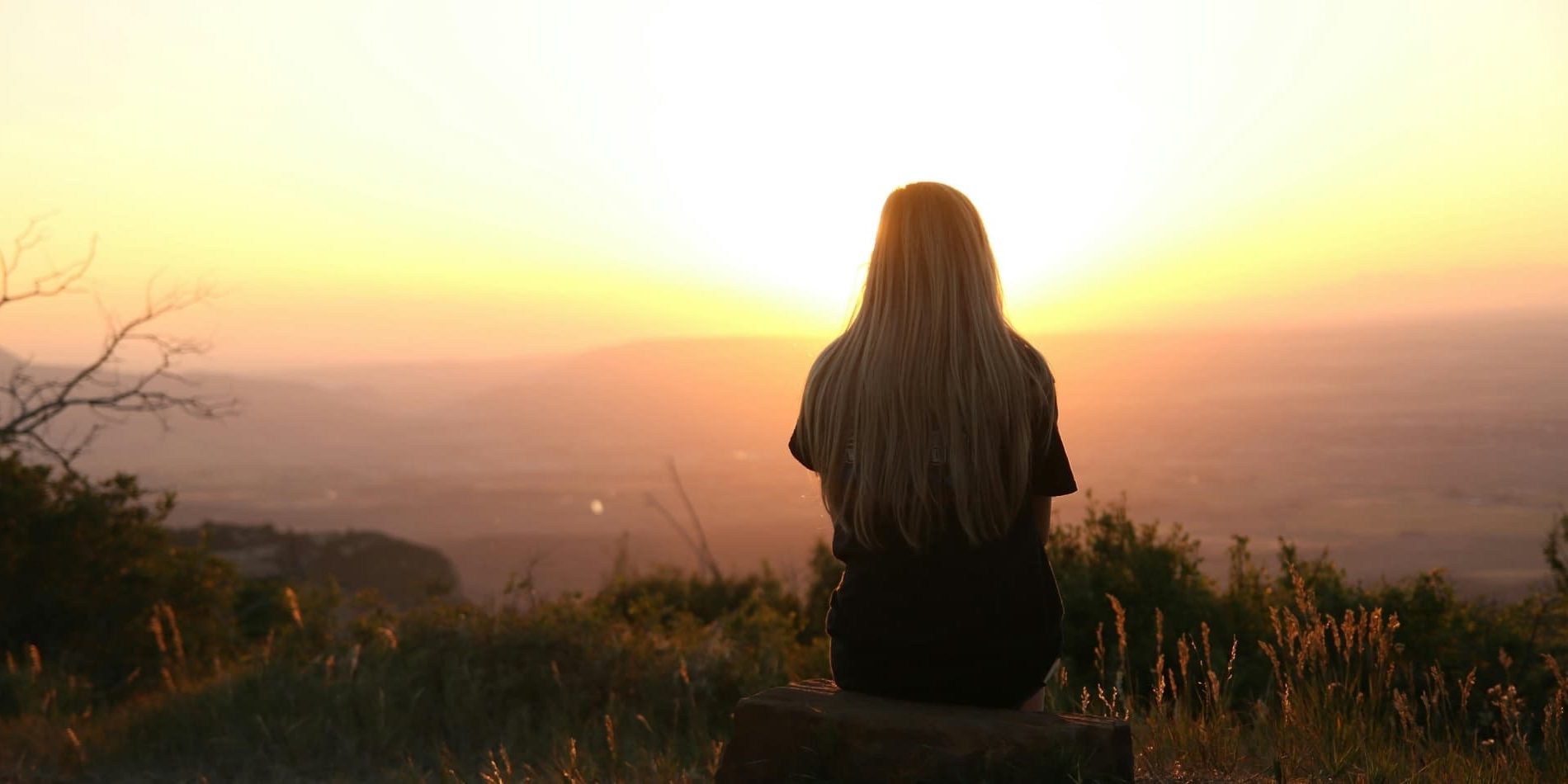 A woman sits alone on a log at sunset, back turned, looking out over a vast landscape