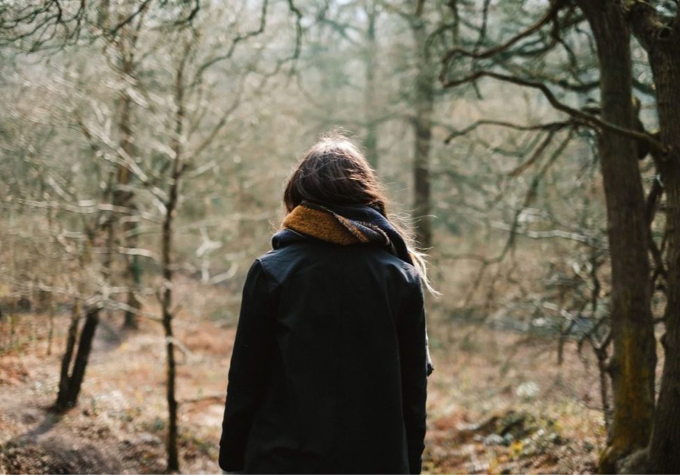 A woman stands alone in a winter forest, her back to the camera, surrounded by bare trees.