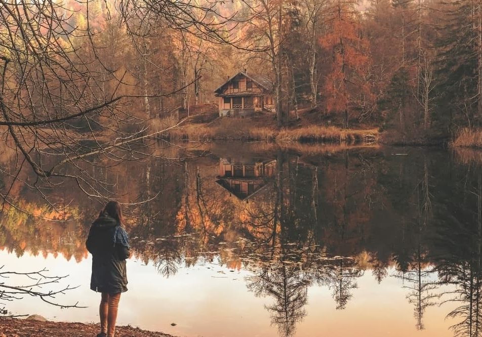 A woman stands at the edge of a still lake reflecting an autumn forest and a house on the far shore